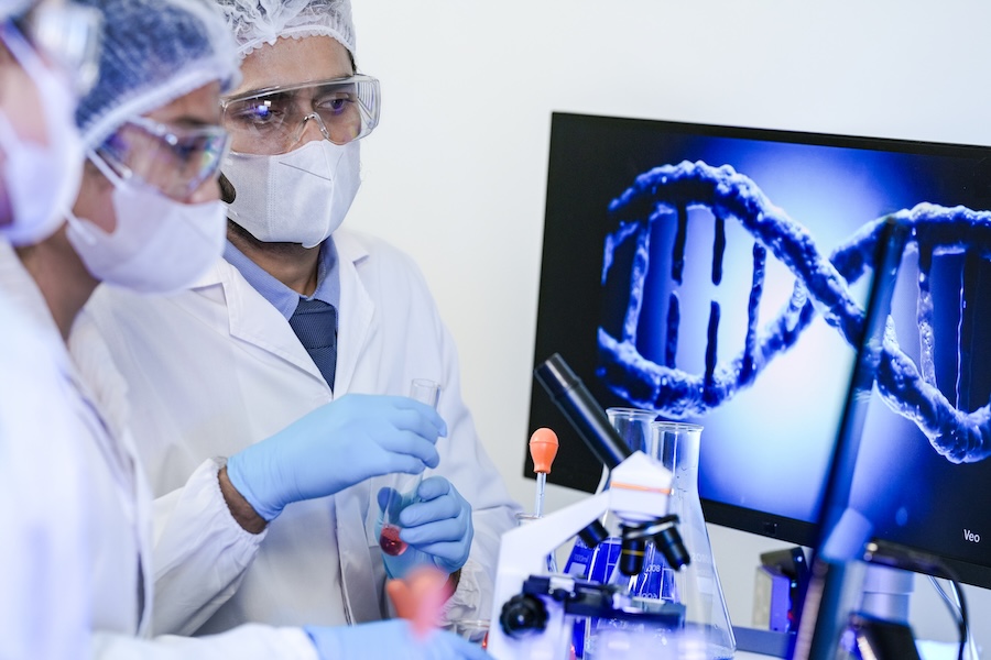 A diverse team of scientists collaborates in a genetics lab. A male researcher explains a procedure with a liquid sample while a DNA helix is displayed on a monitor behind them.